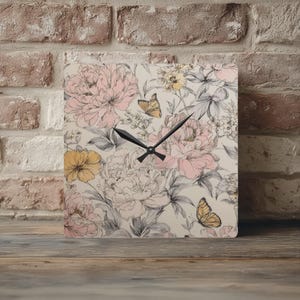 May include: Square wall clock with a floral design featuring pink peonies, yellow flowers, and butterflies on a cream background. The clock hands are black. The clock is sitting on a wooden surface.