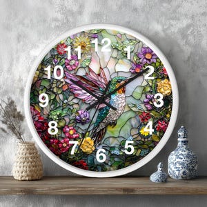 May include: White-framed wall clock featuring a stained-glass design with a hummingbird and floral elements. The clock face displays large white numerals against a backdrop of vibrant colors, including pink, green, and yellow. Black clock hands indicate the time.