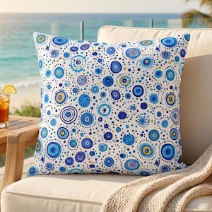May include: White decorative throw pillow with a blue and yellow circular pattern. The pillow is on a beige chair with a tan blanket. A glass of iced tea and a small succulent plant are on a wooden table.