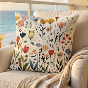 May include: A square decorative pillow with a floral pattern. The pillow features a cream-colored background with colorful flowers in shades of red, yellow, blue, and orange. The pillow is placed on a beige armchair with a knitted throw blanket.