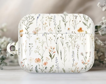 Puede incluir: Funda blanca para AirPods con un delicado estampado floral. El diseño presenta una variedad de flores silvestres en tonos melocotón, lavanda y verde, sobre un fondo blanco. La funda tiene un acabado liso y brillante y un pequeño lazo para sujetar.