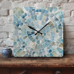Pode incluir: Relógio de parede quadrado com um design de mosaico em tons de azul, branco e bege. O mostrador do relógio apresenta um padrão radial de pequenas telhas irregulares. Os ponteiros pretos indicam as horas. À esquerda, um pequeno vaso decorativo azul e branco.