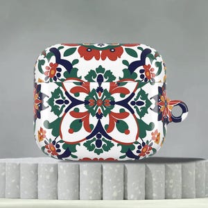 May include: White AirPod case with a colorful floral pattern in red, green, blue, and yellow. The case has a rounded rectangular shape with a small loop on the side. The design is reminiscent of a decorative tile.