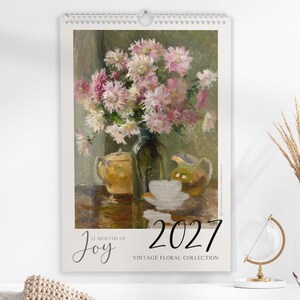Op de afbeelding: Een wandkalender voor 2027 met een stilleven olieverfschilderij van een boeket roze en witte bloemen in een glazen vaas, vergezeld door een gele theepot, een kleine kan en een wit theekopje op een tafel. De tekst luidt 12 Months of Joy 2027.