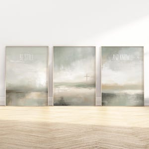 Könnte beinhalten: Drei gerahmte Landschaftskunst-Drucke in neutralen Farbtönen. Die Drucke zeigen abstrakte Landschaften mit den Worten "BE STILL" und "AND KNOW". Ein Kreuz ist auf dem mittleren Druck zu sehen. Das Kunstwerk wird auf einem Holzboden präsentiert.