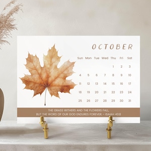 May include: A desktop calendar for October featuring a large, detailed brown maple leaf with water droplets. The calendar has a white background with a brown border and gold-colored metal stand. The text reads "October" and includes the days of the month.