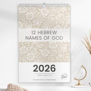 Könnte beinhalten: Wandkalender mit floralem Muster und dem Text "12 Hebräische Namen Gottes" und "2026". Der Kalender hat eine Spiralbindung oben. Eine Vase mit getrockneten Pflanzen und ein Globus befinden sich im Hintergrund.