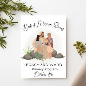 May include: A white card featuring the text "Book of Mormon Stories" and an illustration of figures. Below, it reads "LEGACY 3RD WARD Primary Program October 8th." Greenery and a beige cloth are in the background.