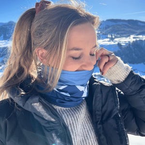Könnte beinhalten: Ein blau-weiß gemusterter Schlauchschal. Der Schlauchschal wird in einer schneebedeckten Berglandschaft getragen. Die Person trägt eine schwarze Jacke und einen grauen Pullover. Der Himmel ist klar und blau.