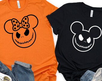 Puede incluir: Dos camisetas con temática de Halloween. Una es naranja con una cabeza de Mickey Mouse en blanco y negro con la cara de Jack Skellington. La otra es negra con una cabeza de Mickey Mouse en blanco con la cara de Jack Skellington.