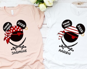 May include: Two white t-shirts with a black Mickey Mouse head wearing a red and white pirate bandana and an eye patch. The text "Disney Magic" and "2025" are on the shirts. The names "Jasmine" and "Aaron" are printed on the shirts.