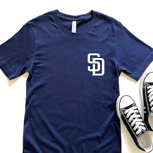 May include: Navy blue t-shirt with a white "SD" logo and a pair of black and white sneakers. The t-shirt is short-sleeved and has a crew neck. The sneakers are low-top and have white laces.