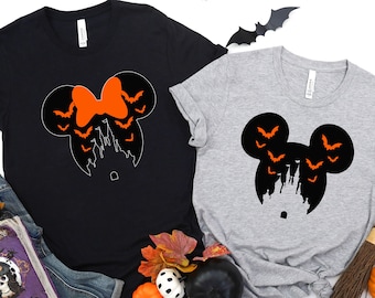 May include: Two black t-shirts with a Halloween-themed Mickey Mouse design. One shirt features a black Mickey Mouse head with orange bats and a castle inside the ears. The other shirt features a black Minnie Mouse head with a red bow and orange bats and a castle inside the ears. The shirts are laid out on a white surface with a book, pumpkins, and a broom.