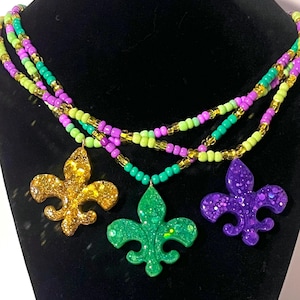 Puede incluir: Un collar de cuentas de varias hebras con tres colgantes de flor de lis en acabados de purpurina dorada, verde y morada. El collar está compuesto por pequeñas cuentas redondas en tonos alternos de verde lima, morado brillante y oro translúcido.
