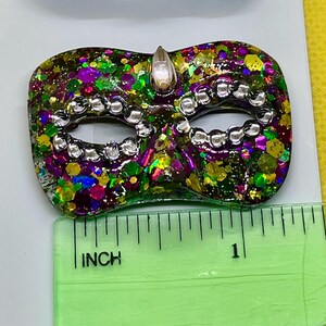 Puede incluir: Un broche con forma de m&aacute;scara de carnaval con un acabado de purpurina multicolor en tonos verde, morado y dorado, acentuado con cuentas plateadas alrededor de los ojos y una gema transparente en la parte superior, de unos 3,8 cm de ancho.