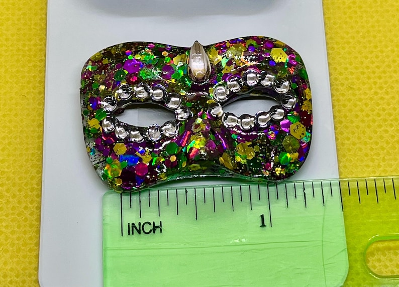 Puede incluir: Un broche con forma de m&aacute;scara de carnaval con un acabado de purpurina multicolor en tonos verde, morado y dorado, acentuado con cuentas plateadas alrededor de los ojos y una gema transparente en la parte superior, de unos 3,8 cm de ancho.