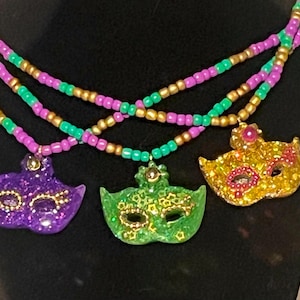 Puede incluir: Un collar de varias hebras con tres colgantes en forma de máscara en colores morado, verde y dorado. Las cuentas están dispuestas en patrones alternos de morado, verde y oro metálico, creando un aspecto vibrante y superpuesto con acabados brillantes.