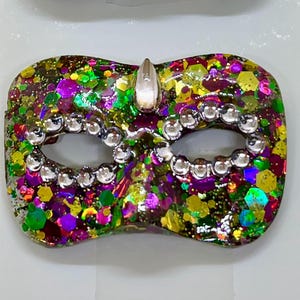 Puede incluir: Una m&aacute;scara de carnaval con una mezcla vibrante de purpurina hexagonal morada, verde y dorada incrustada en una base de resina transparente. Las aberturas de los ojos est&aacute;n delineadas con peque&ntilde;os diamantes de imitaci&oacute;n redondos y una gema en forma de l&aacute;grima en el centro.