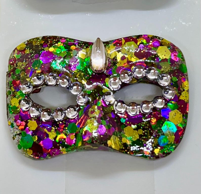 Puede incluir: Una m&aacute;scara de carnaval con una mezcla vibrante de purpurina hexagonal morada, verde y dorada incrustada en una base de resina transparente. Las aberturas de los ojos est&aacute;n delineadas con peque&ntilde;os diamantes de imitaci&oacute;n redondos y una gema en forma de l&aacute;grima en el centro.
