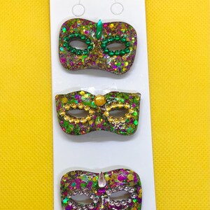 Puede incluir: Tres broches decorativos de m&aacute;scara de carnaval sujetos a una tarjeta de presentaci&oacute;n blanca. Cada m&aacute;scara tiene un acabado brillante en morado, verde y dorado. La m&aacute;scara superior tiene cuentas y una gema verdes, la del medio cuentas y una gema doradas, y la inferior cuentas plateadas y una gema transparente.