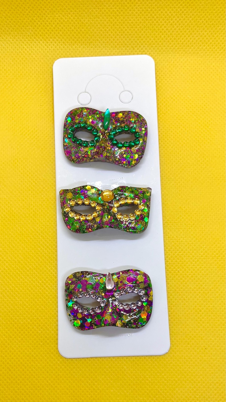 Puede incluir: Tres broches decorativos de m&aacute;scara de carnaval sujetos a una tarjeta de presentaci&oacute;n blanca. Cada m&aacute;scara tiene un acabado brillante en morado, verde y dorado. La m&aacute;scara superior tiene cuentas y una gema verdes, la del medio cuentas y una gema doradas, y la inferior cuentas plateadas y una gema transparente.