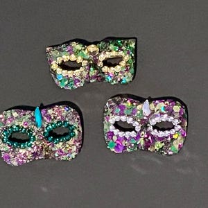 Puede incluir: Tres m&aacute;scaras de carnaval decorativas con una superficie texturizada cubierta de peque&ntilde;os adornos brillantes en tonos morado, verde y dorado. Cada m&aacute;scara tiene dos aberturas para los ojos y est&aacute; decorada con peque&ntilde;as cuentas o cristales.
