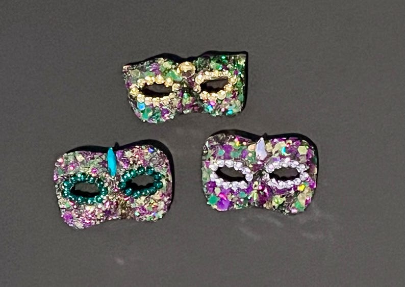 Puede incluir: Tres m&aacute;scaras de carnaval decorativas con una superficie texturizada cubierta de peque&ntilde;os adornos brillantes en tonos morado, verde y dorado. Cada m&aacute;scara tiene dos aberturas para los ojos y est&aacute; decorada con peque&ntilde;as cuentas o cristales.