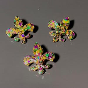 Puede incluir: Tres adornos con forma de flor de lis que presentan una base de resina transparente incrustada con una densa mezcla de purpurina dorada, morada y verde. Cada pieza muestra un diseño clásico de tres pétalos con una punta central.