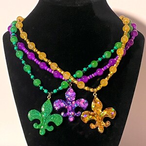 Puede incluir: Un collar de cuentas de varias hebras con tres colgantes de flor de lis en verde, morado y dorado. Las cuentas están dispuestas en patrones alternos de verde, morado y dorado, con cada colgante cubierto de purpurina y lentejuelas a juego.