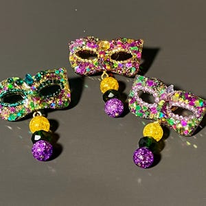 Puede incluir: Tres pendientes con forma de máscara de Mardi Gras con un acabado brillante en morado, verde y dorado. Cada máscara está adornada con un dije colgante que consta de tres cuentas esféricas en colores morado, verde y dorado a juego.