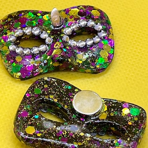Puede incluir: Dos objetos con forma de m&aacute;scara de carnaval con una base oscura llena de purpurina morada, verde y dorada. Una m&aacute;scara presenta pedrer&iacute;a plateada alrededor de los ojos, mientras que la otra tiene un im&aacute;n circular plateado en la parte posterior.