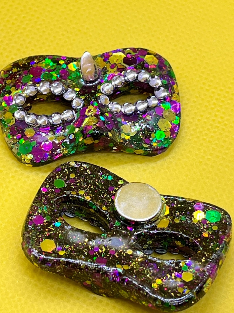 Puede incluir: Dos objetos con forma de m&aacute;scara de carnaval con una base oscura llena de purpurina morada, verde y dorada. Una m&aacute;scara presenta pedrer&iacute;a plateada alrededor de los ojos, mientras que la otra tiene un im&aacute;n circular plateado en la parte posterior.