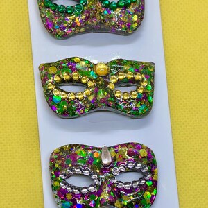 Puede incluir: Tres broches de m&aacute;scara de carnaval sujetos a una tarjeta blanca. Cada m&aacute;scara presenta un acabado de purpurina multicolor en tonos verde, morado y dorado, con cuentas met&aacute;licas alrededor de los ojos y una gema en la parte superior central.