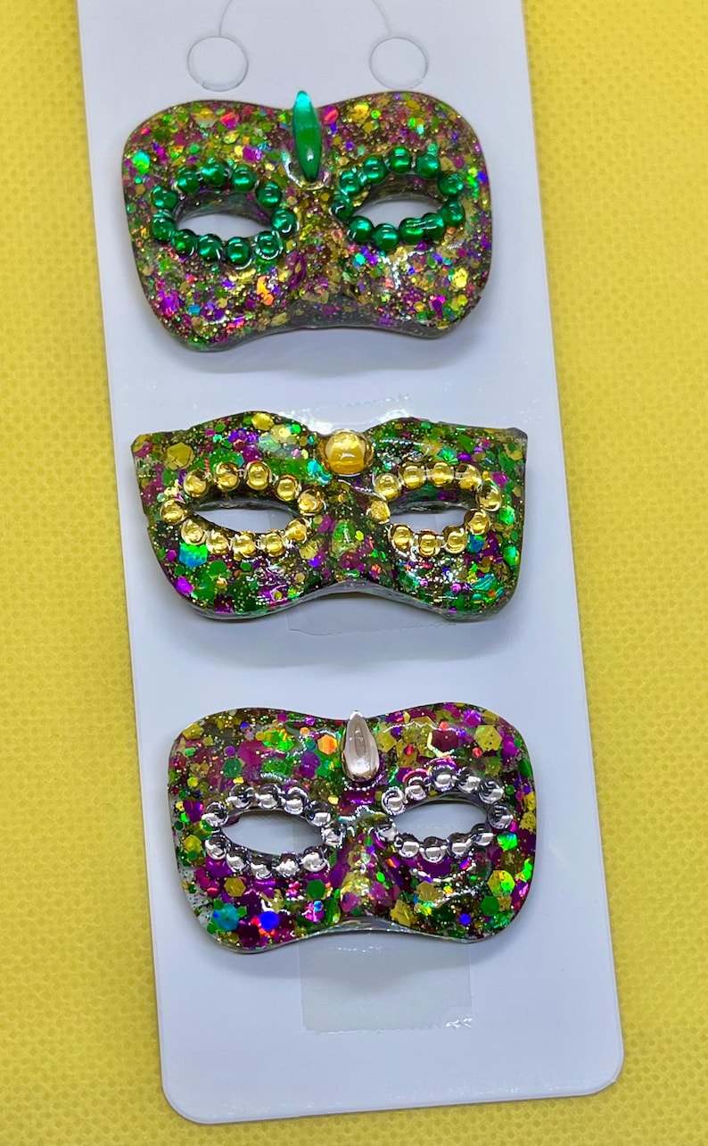 Puede incluir: Tres broches de m&aacute;scara de carnaval sujetos a una tarjeta blanca. Cada m&aacute;scara presenta un acabado de purpurina multicolor en tonos verde, morado y dorado, con cuentas met&aacute;licas alrededor de los ojos y una gema en la parte superior central.