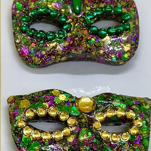 Puede incluir: Dos m&aacute;scaras de carnaval con acabado brillante en tonos verde, morado y dorado. La m&aacute;scara superior est&aacute; decorada con cuentas verdes y una gema verde, mientras que la inferior tiene cuentas doradas y una gema circular central de tono dorado.