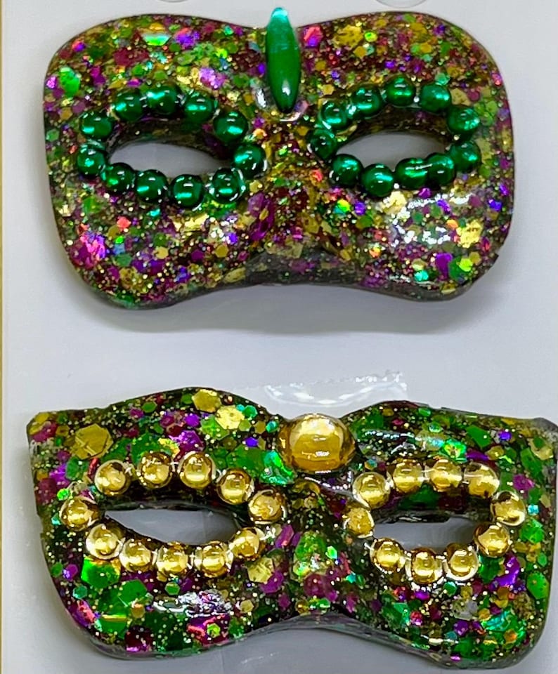 Puede incluir: Dos m&aacute;scaras de carnaval con acabado brillante en tonos verde, morado y dorado. La m&aacute;scara superior est&aacute; decorada con cuentas verdes y una gema verde, mientras que la inferior tiene cuentas doradas y una gema circular central de tono dorado.