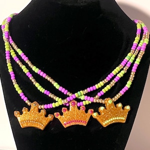Puede incluir: Un collar de cuentas de varias hebras con tres colgantes de corona con purpurina dorada. El collar está compuesto por pequeñas cuentas redondas en colores alternos de verde lima brillante, morado vibrante y oro metálico en capas.
