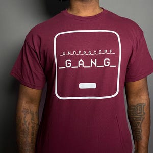 May include: A maroon short-sleeved t-shirt featuring a white graphic print on the chest. The design consists of a rounded square outline containing the text _U_N_D_E_R_S_C_O_R_E_ and _G_A_N_G_ in a pixelated font, with a horizontal underscore symbol below.