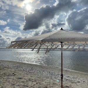 May include: A white beach umbrella with a fringed border stands on a sandy beach with a body of water in the background. The sun is shining brightly in the sky, which is filled with fluffy white clouds.