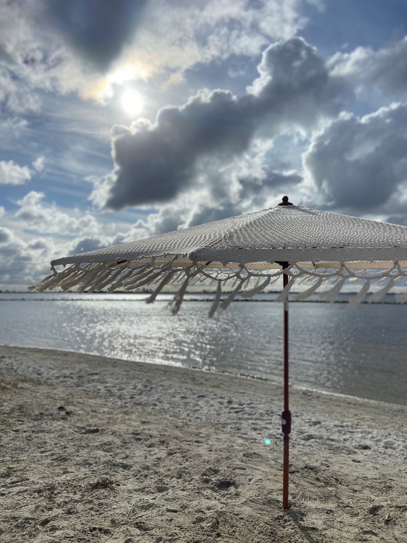 May include: A white beach umbrella with a fringed border stands on a sandy beach with a body of water in the background. The sun is shining brightly in the sky, which is filled with fluffy white clouds.