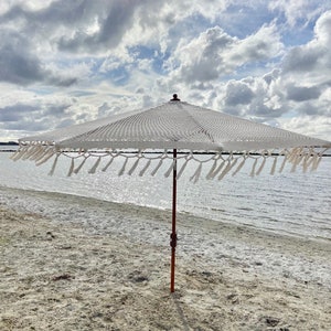 May include: White beach umbrella with a wooden pole and fringe trim. The umbrella is on a sandy beach with a body of water in the background.