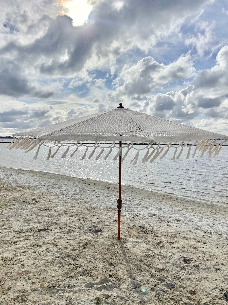 May include: White beach umbrella with a wooden pole and fringe trim. The umbrella is on a sandy beach with a body of water in the background.