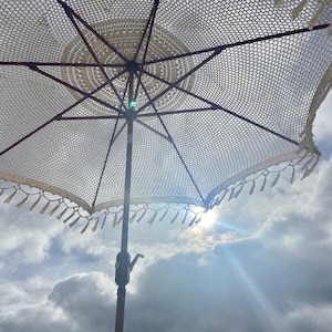 May include: A white crocheted beach umbrella with tassels hanging from the edge. The umbrella is open and the sun is shining through the fabric.
