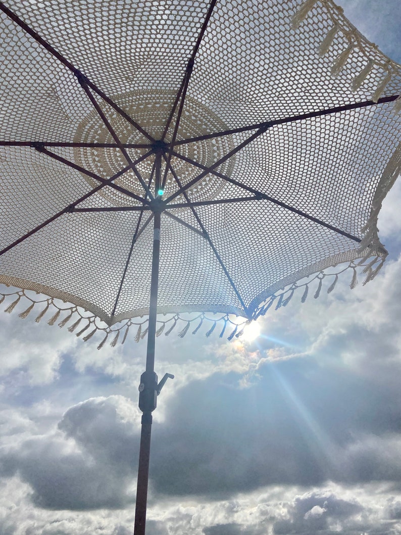 May include: A white crocheted beach umbrella with tassels hanging from the edge. The umbrella is open and the sun is shining through the fabric.