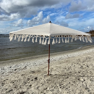 May include: A white crocheted beach umbrella with a wooden pole and fringe trim. The umbrella is on a sandy beach with a body of water in the background.