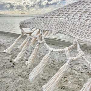 May include: White cotton hammock with tassels hanging from the edge. The hammock is hanging over a sandy beach with a cloudy sky in the background.