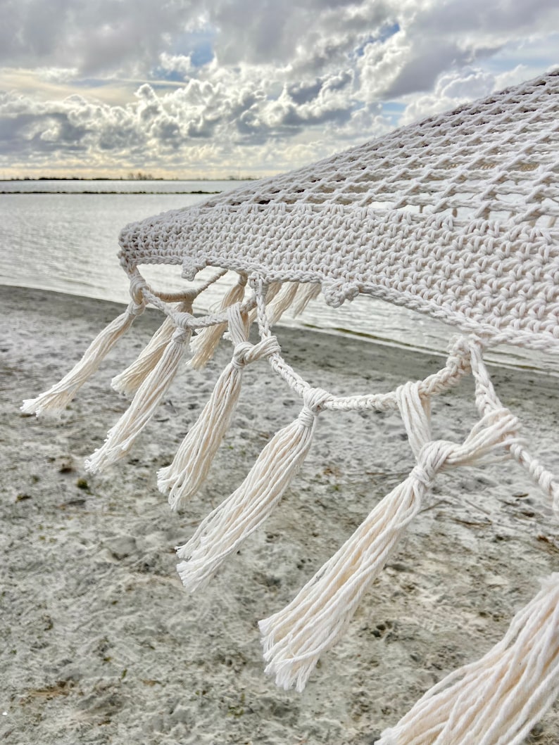 May include: White cotton hammock with tassels hanging from the edge. The hammock is hanging over a sandy beach with a cloudy sky in the background.