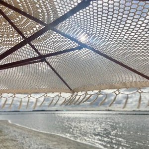May include: A white crocheted beach umbrella with fringe hanging over a sandy beach and a body of water in the background.