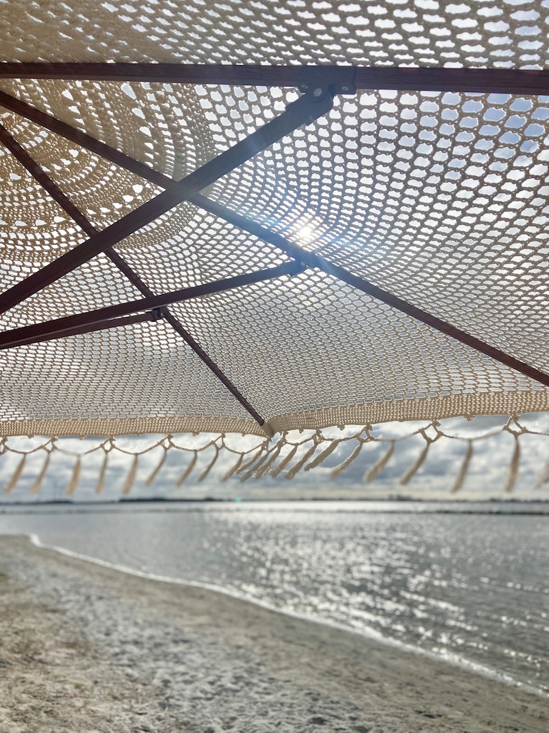May include: A white crocheted beach umbrella with fringe hanging over a sandy beach and a body of water in the background.