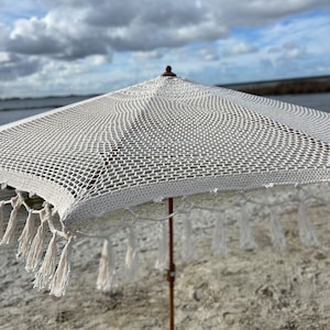 May include: A white crochet beach umbrella with a wooden pole and tassels hanging from the edge. The umbrella is set up on a sandy beach.